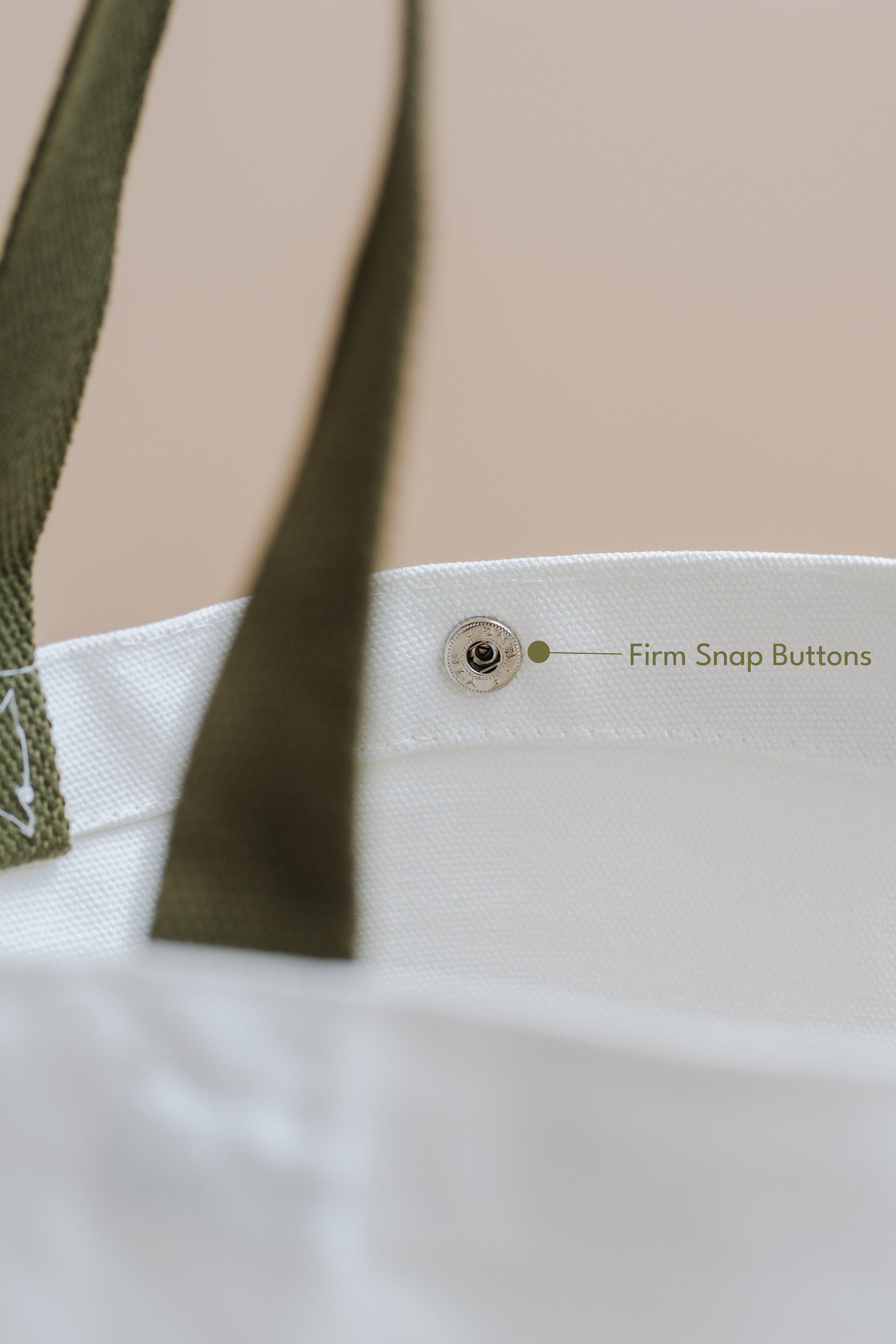 Close-up of a white tote bag with a green strap and snap button, on a beige background.