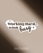 Sticker with text 'Working Hard... to look busy' on a textured beige background